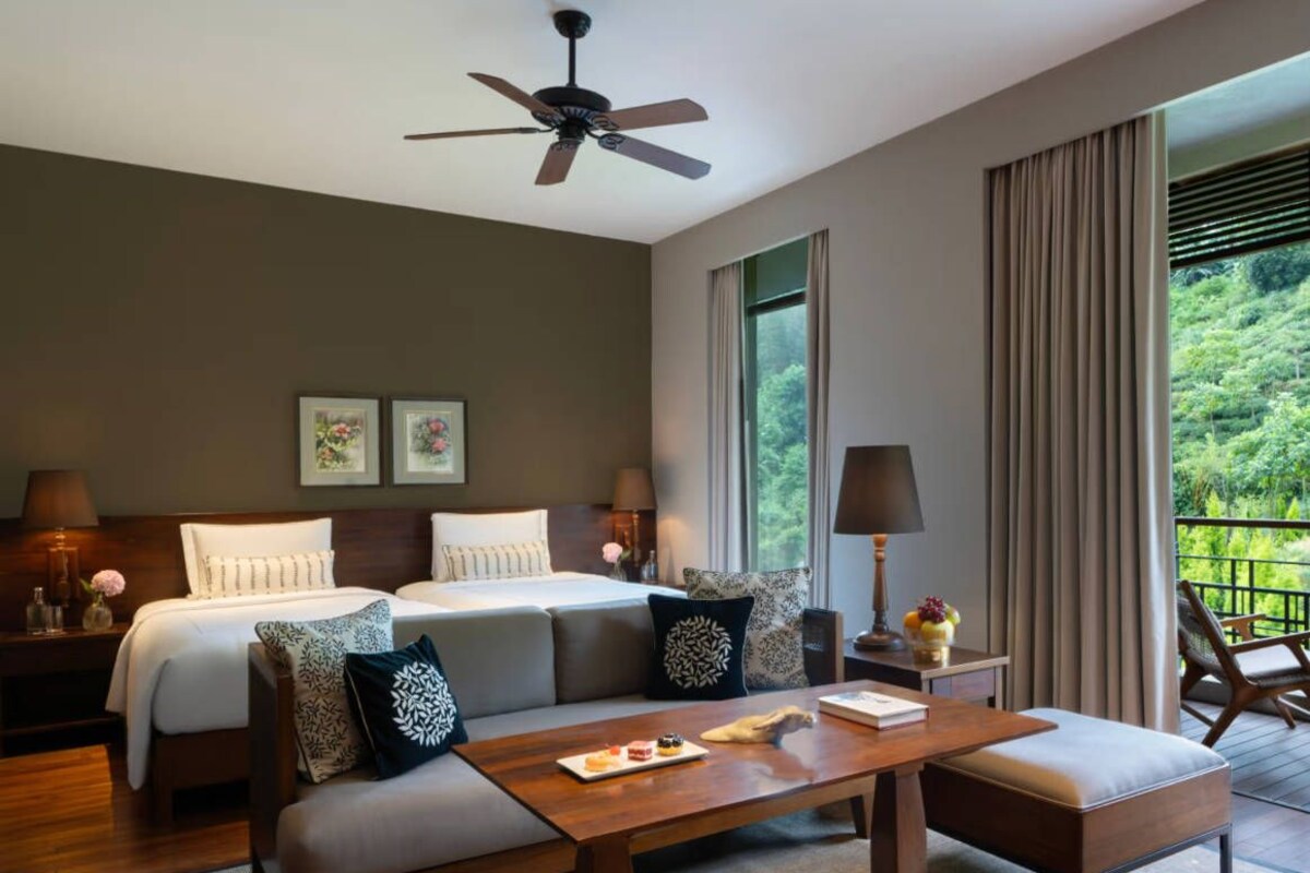 A hotel room with a bed, a sofa, a table with a lamp, and a view of greenery outside.