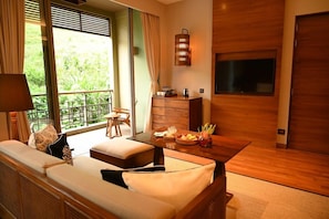 A room with a sofa, a dining table with fruit, a TV, and a view of greenery outside.
