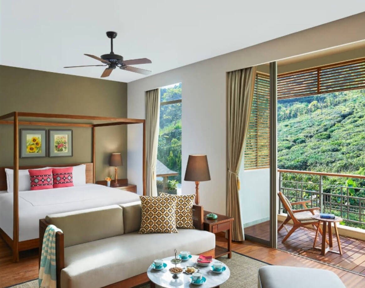 A hotel room with a four-poster bed, a sofa, a balcony with a view of a lush green landscape, and a ceiling fan.