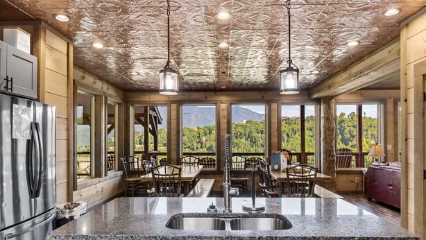 Una cocina con encimeras de granito, electrodomésticos de acero inoxidable y una isla grande con un lavabo. Hay sillas y mesas de madera, y ventanas amplias que ofrecen vistas de un paisaje montañoso.