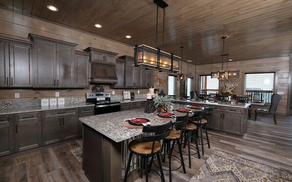 A spacious kitchen with granite countertops, dark wood cabinets, and a central island with bar stools. - Cavern Cascades - Most unique pool in the Smokies! Luxury Interior, Cozy Hot Tub & Stunning Views (Sevierville)