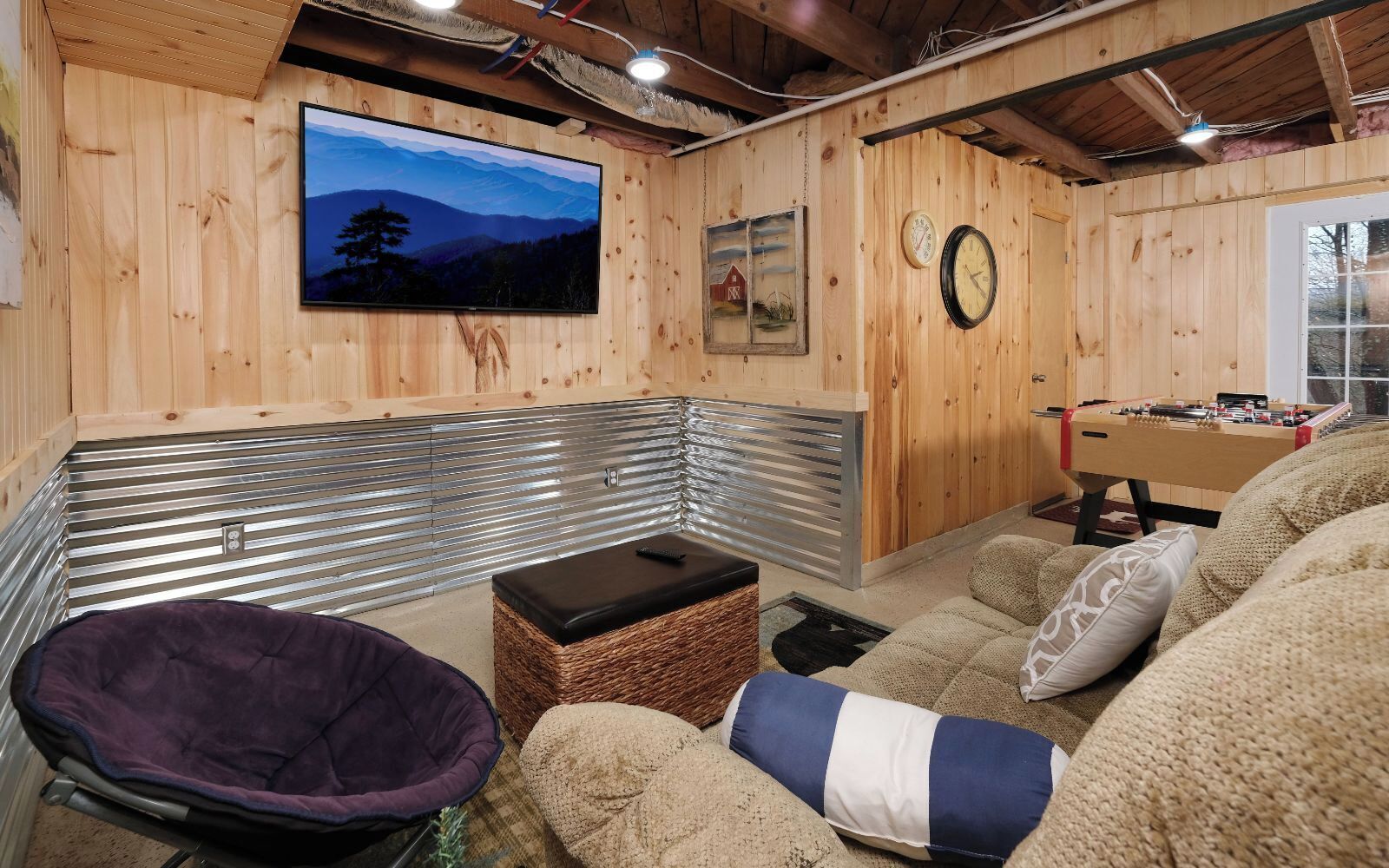 Um quarto aconchegante com decoração em madeira, uma televisão montada na parede, uma mesa de pebolim e uma área de estar confortável com poltrona e sofá estofados.