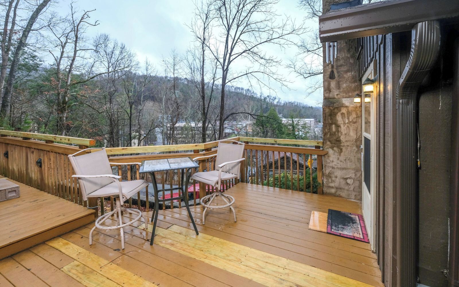 A wooden deck with outdoor furniture, a stone wall, and a view of trees and hills.