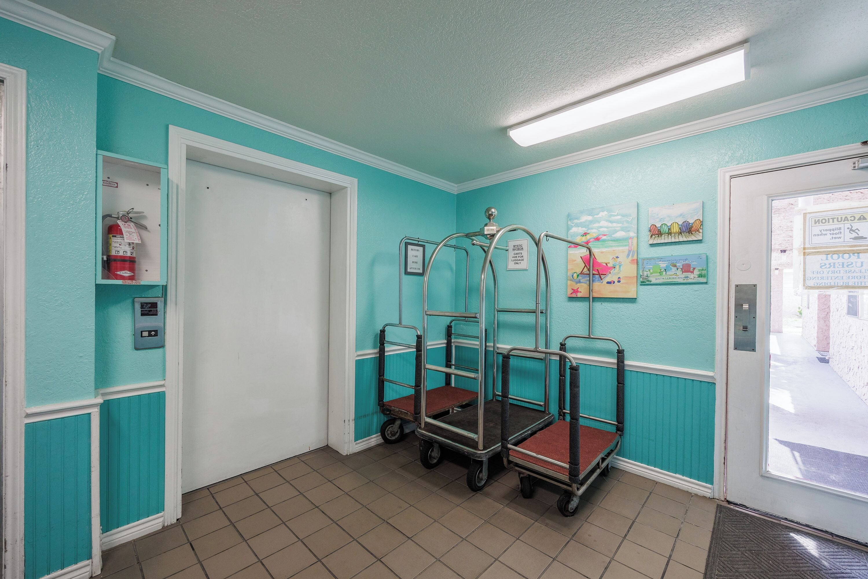 A teal-walled room with a white door, a fire extinguisher, and a cart with shelves.