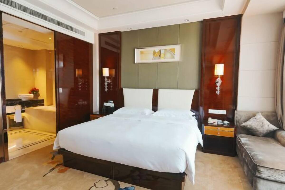 A hotel room with a large bed, a sofa, a desk with a lamp, and a painting on the wall.