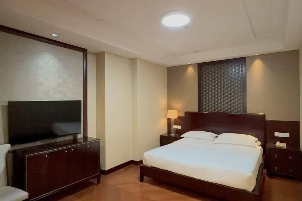 A hotel room with a bed, a TV, a nightstand, and a wall-mounted light fixture.