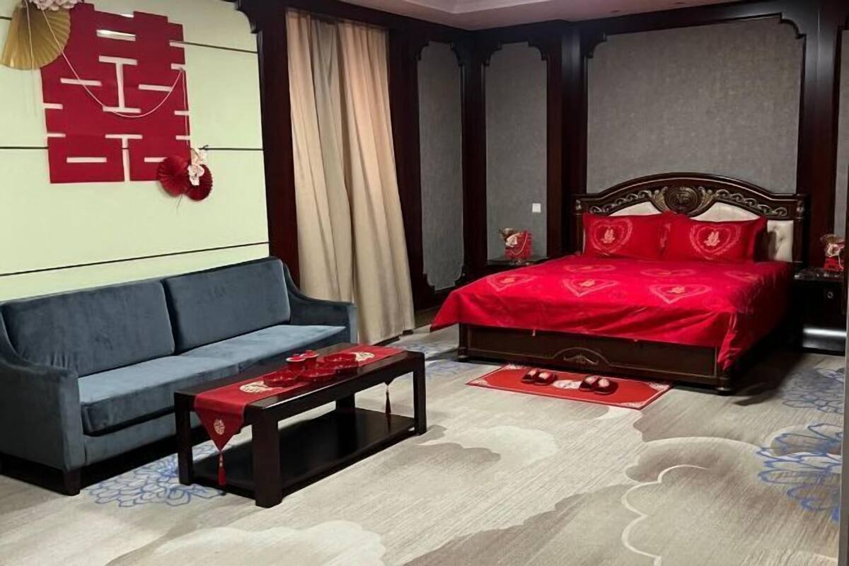 Quarto com sofá, cama com roupa de cama vermelha, mesa pequena e um objeto decorativo na parede.