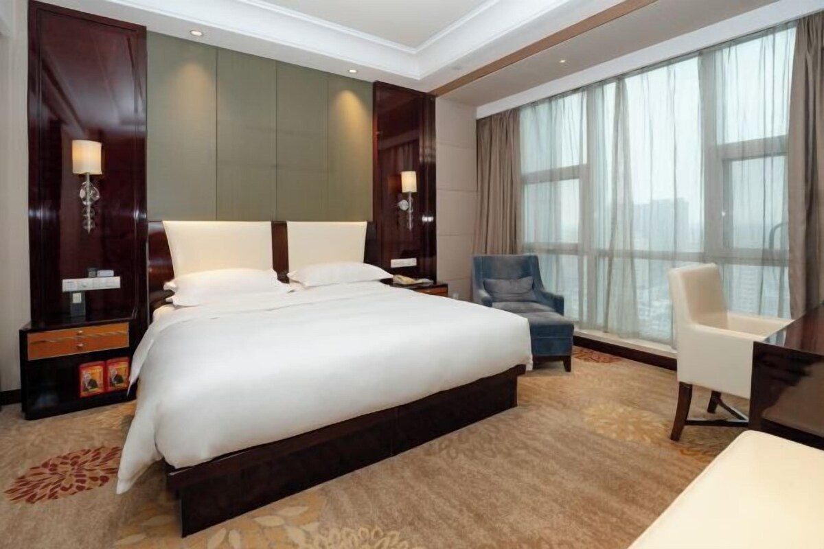A hotel room with a large bed, a desk, a chair, and a large window.