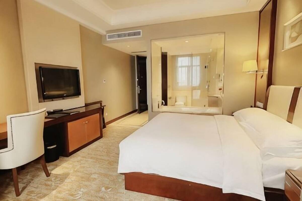 A hotel room with a bed, a desk with a TV, a chair, a bathroom with a mirror, and a window.