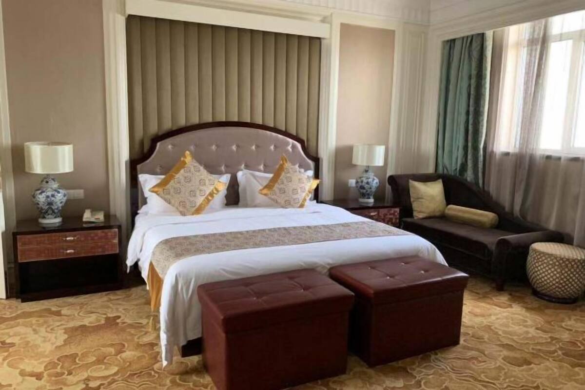 Quarto de hotel com uma cama grande, duas mesas de cabeceira, um sofá e um tapete estampado.