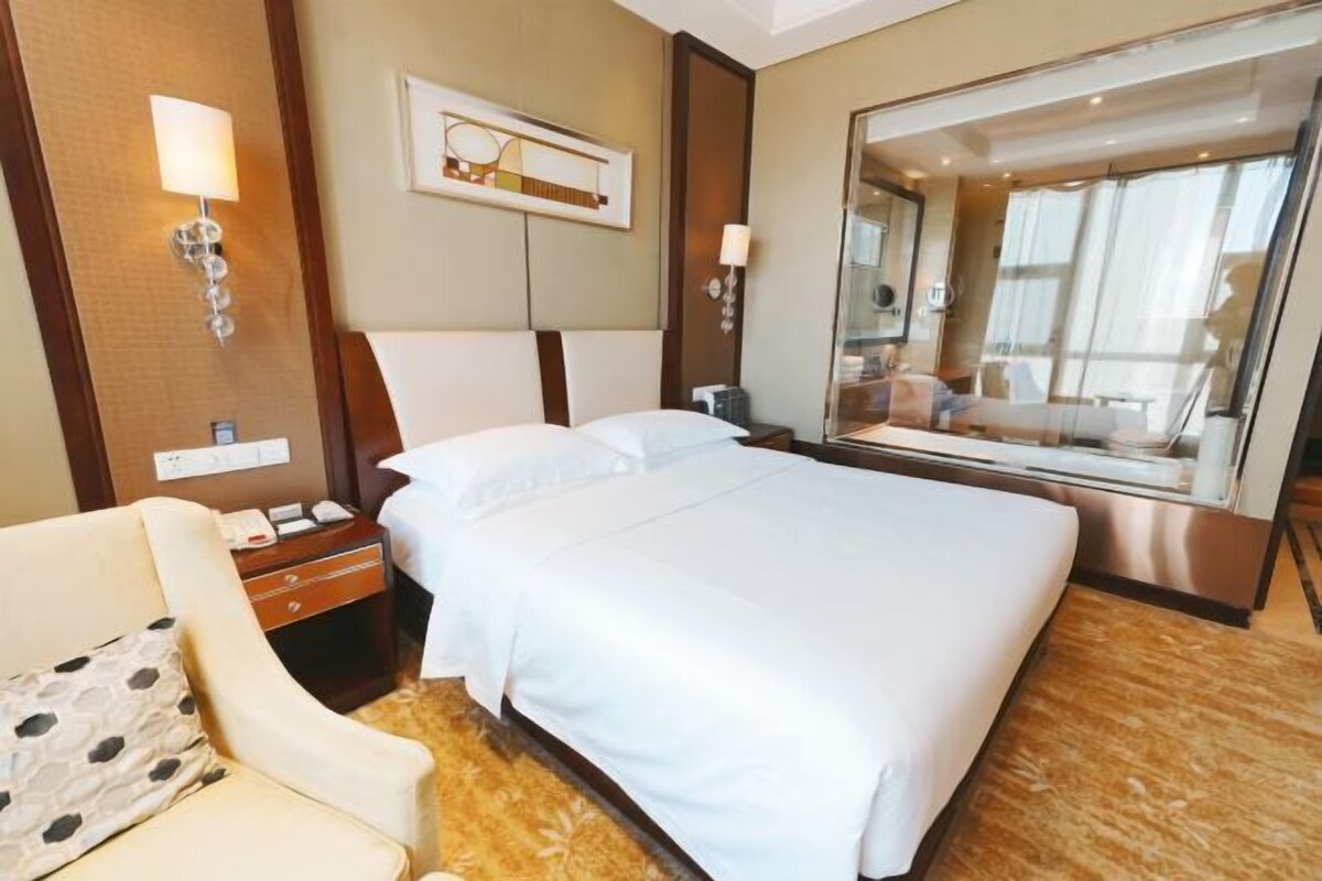 A hotel room with a large bed, a bedside table with a lamp, a chair, and a large mirror reflecting the room.