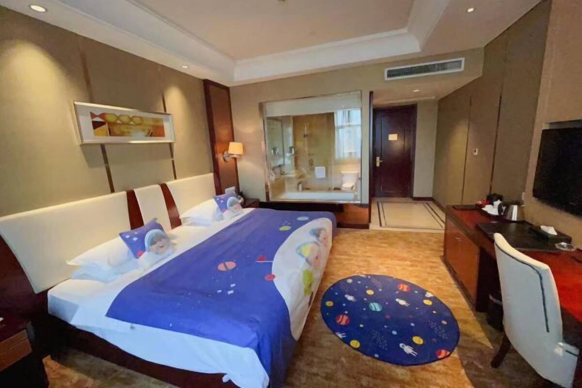 A hotel room with a large bed, a desk, a chair, a TV, and a bathroom visible through a glass door.