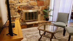 Ein Wohnzimmer mit Kamin aus Stein, einem Glastisch, einem Fernsehschrank aus Holz und einem grauen Sessel.