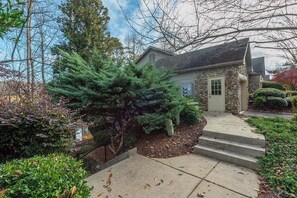 Ein Steinhaus mit Veranda, Stufen zum Eingang und einem gepflegten Garten mit verschiedenen Sträuchern und einem großen Nadelbaum.