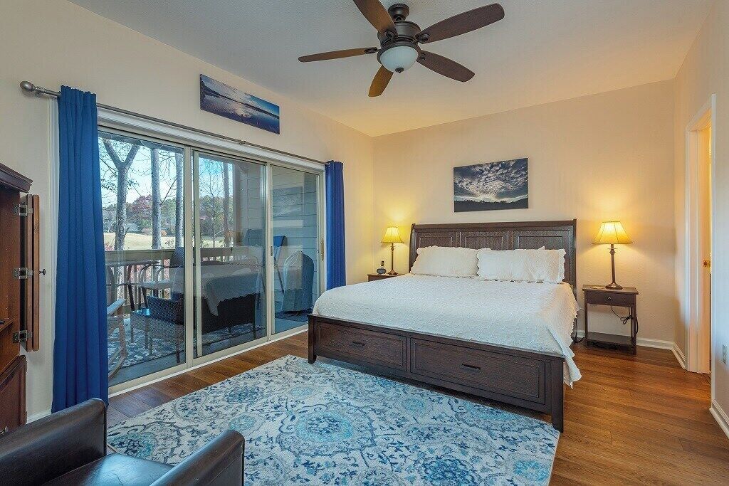 A bedroom with a large bed, two bedside lamps, a ceiling fan, a sliding glass door to a balcony, and a view of trees.