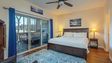 A bedroom with a large bed, two bedside lamps, a ceiling fan, a sliding glass door to a balcony, and a view of trees.
