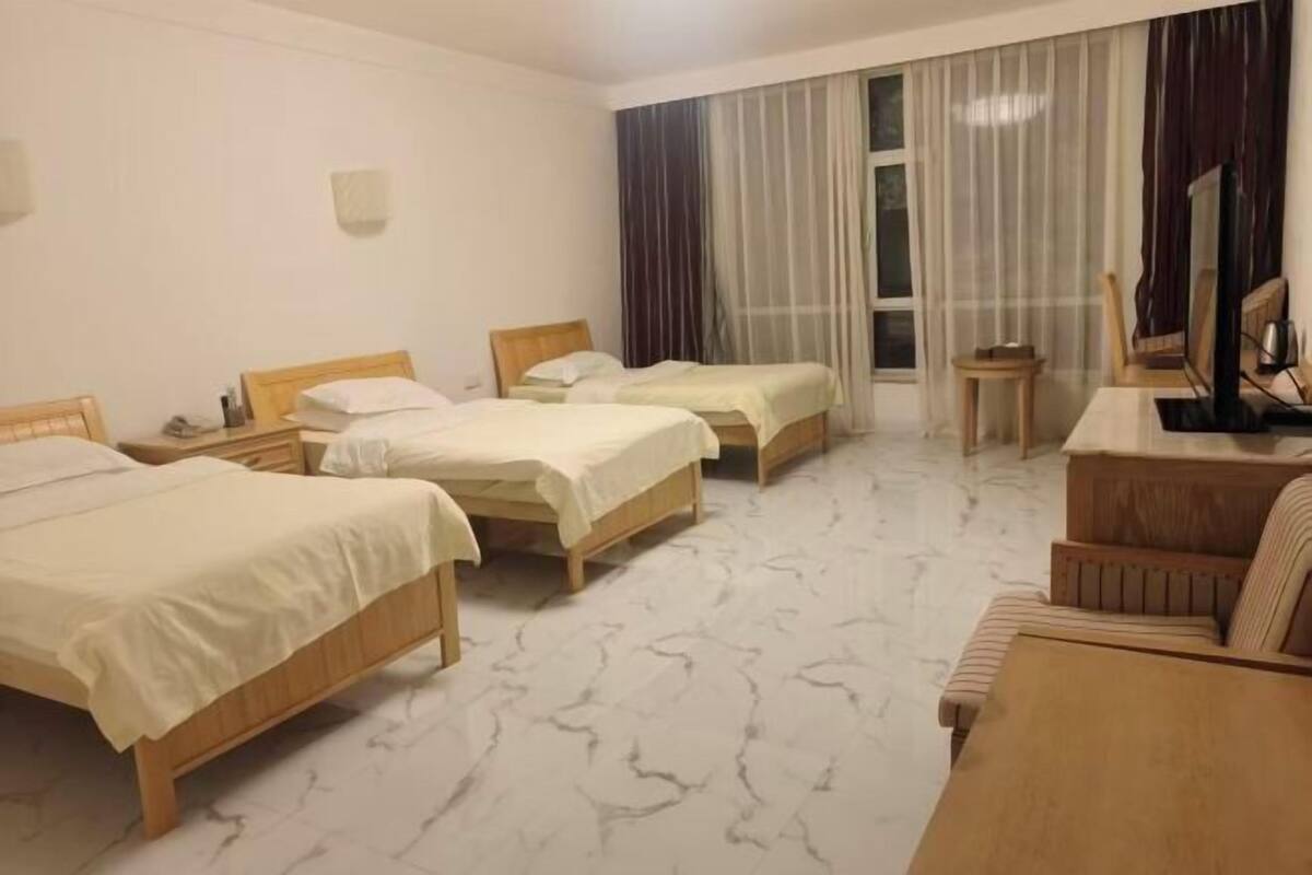 A hotel room with three beds, a wooden desk, a chair, and a large window with curtains.