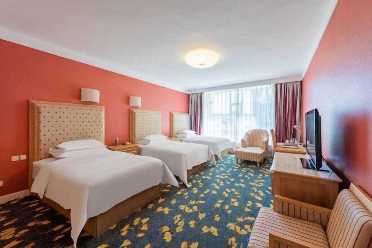 Ein Hotelzimmer mit drei Betten, einem Sofa, einem Fernseher und einem Fenster mit Vorhängen.