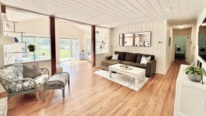 Uma sala de estar ampla com piso de madeira, um sofá marrom, uma mesa de centro branca e uma poltrona estampada.