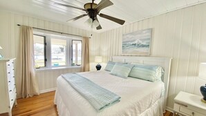 Quarto com uma cama grande, um ventilador de teto, janela com cortinas, mesa de cabeceira e um quadro na parede.