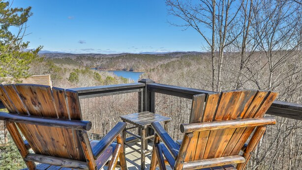 Eine Holzterrasse mit zwei Adirondack-Stühlen und einem kleinen Tisch, mit Blick auf einen See und bewaldete Hügel.
