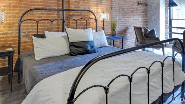 A room with exposed brick walls, a metal bed frame, a small bedside table, and a hanging light fixture. - Elle & Beck`s 2 story suite (Galena)