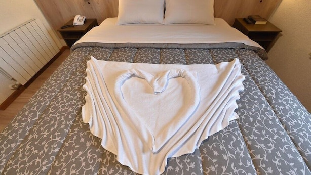 A neatly made bed with a heart-shaped towel arrangement, wooden headboard, bedside lamps, and a telephone on the nightstand. - Hotel Villa Aconchego de Gramado (Gramado)