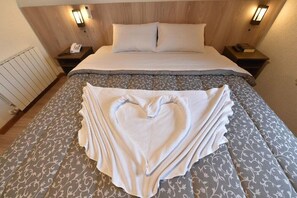 Een net opgemaakt bed met een handdoek in de vorm van een hart, een houten hoofdbord, bedlampjes en een telefoon op het nachtkastje.