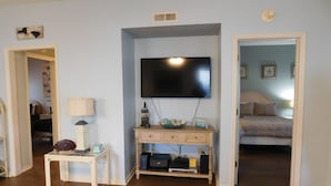 Un saló amb televisió de pantalla plana penjada a la paret, una petita taula de nit de fusta amb una làmpada, i un llit en una habitació adjacent.