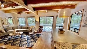 A spacious living room with a large window, a wooden ceiling, and a dining area with white chairs. - Hot tub,pool table,xc/downhill ski,near-Stevens Pass,Leavenworth,Lake Wenatchee (Leavenworth)