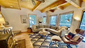 A cozy living room with a fireplace, a sofa, a patterned rug, and large windows offering a view of the outdoors. - Hot tub,pool table,xc/downhill ski,near-Stevens Pass,Leavenworth,Lake Wenatchee (Leavenworth)