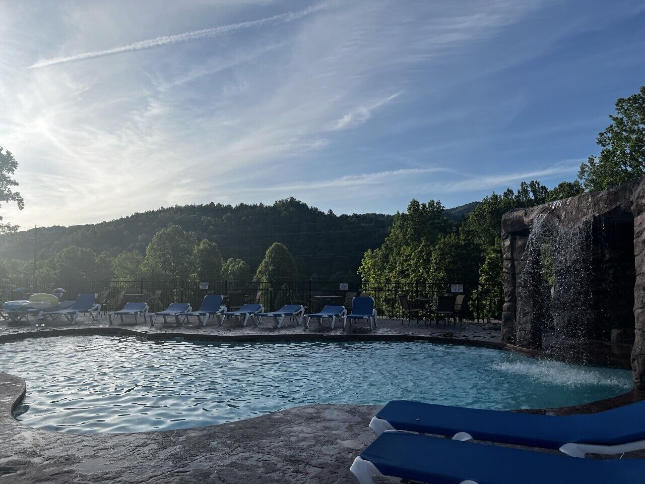 A swimming pool with lounge chairs, a waterfall feature, and a forested hillside in the background.