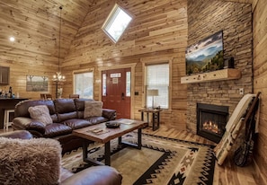 L’intérieur d’une cabane en rondins, avec une cheminée, un canapé en cuir, une table à manger et une vue sur l’extérieur grâce à une lucarne.