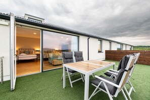 Un patio exterior moderno con una mesa y sillas de madera, un ventanal corredero que da a un espacio interior y vistas del paisaje circundante.