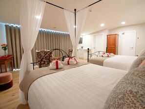 Habitación de hotel con dos camas, una zona de estar con una silla, una mesita con un florero y una puerta de madera.