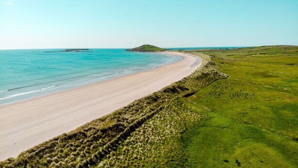 Un paisaje costero con una playa arenosa, campos cubiertos de hierba y una colina al fondo.