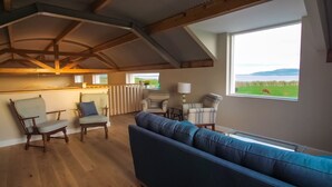 Amplia sala de estar con techo alto, vigas de madera, un ventanal con vista a un campo verde y una vaca, y un sofá azul.