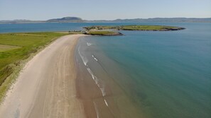 Vista aérea de una zona costera con una extensa playa arenosa, una pequeña isla con edificios y montañas en la distancia.