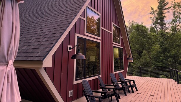 Una cabaña roja de madera con techo inclinado, ventanas grandes y una terraza con sillas.