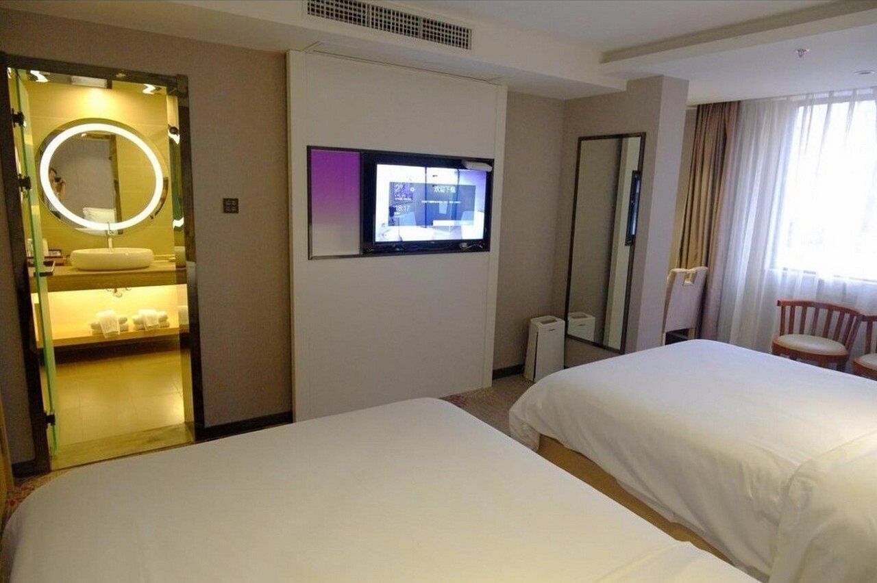 A hotel room with two beds, a bathroom with a mirror and sink, a TV on the wall, and a window with curtains.