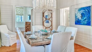 A dining area with a wooden table set for four, surrounded by white chairs. A modern chandelier hangs above the table. - Charming WaterColor Home: Bikes, 6 Seater Electric Cart, Outdoor Fireplace, Steps to Camp WC! (Santa Rosa Beach)