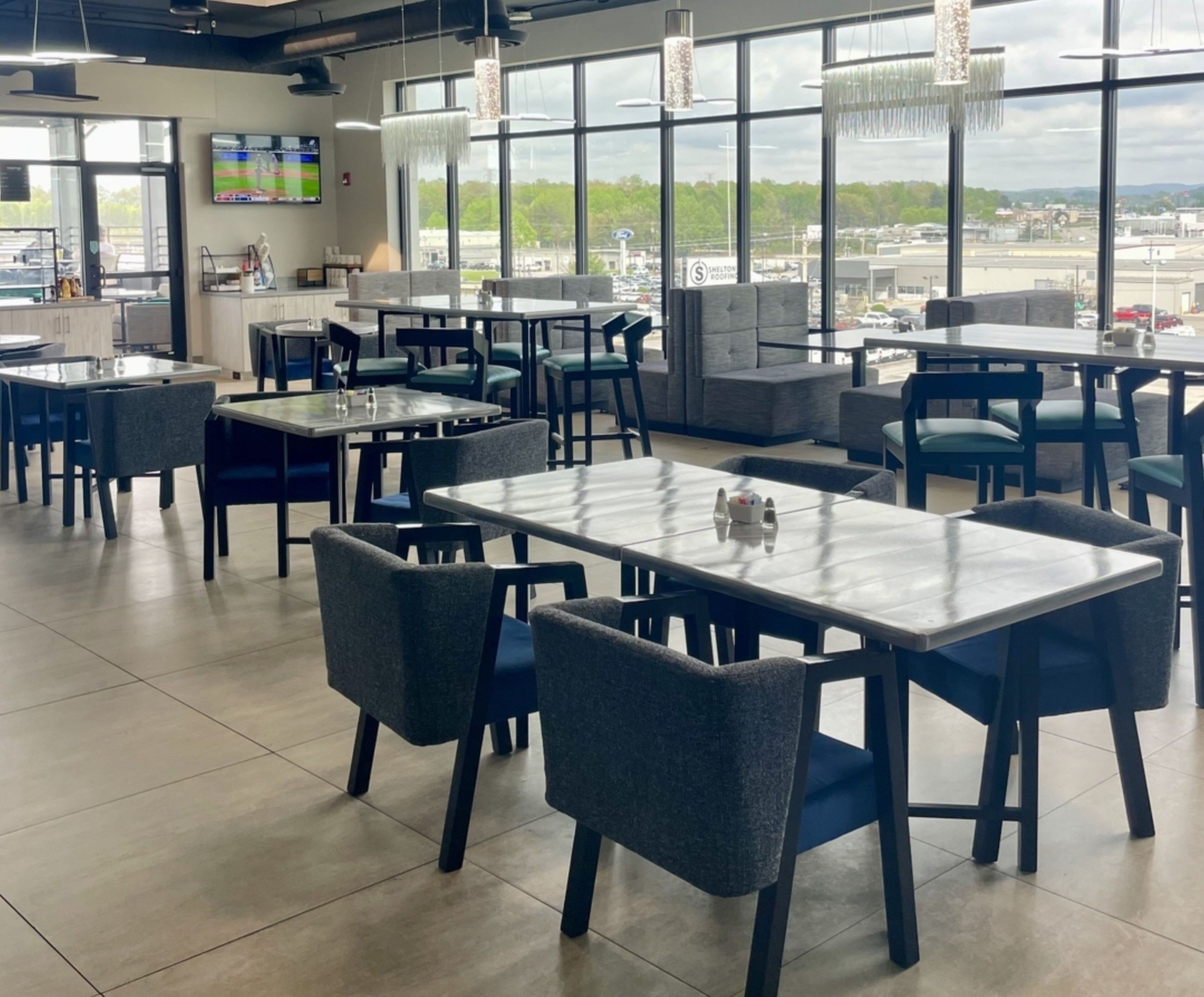 Un interior de restaurante moderno con varias mesas y sillas, ventanas amplias que ofrecen vistas al exterior, y un televisor de pantalla plana montado en la pared.
