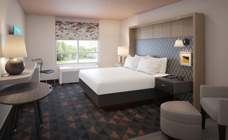 Habitación de hotel con una cama grande, un escritorio, una silla, una mesita pequeña, una mesita de noche con lámpara y una ventana con vista a árboles.