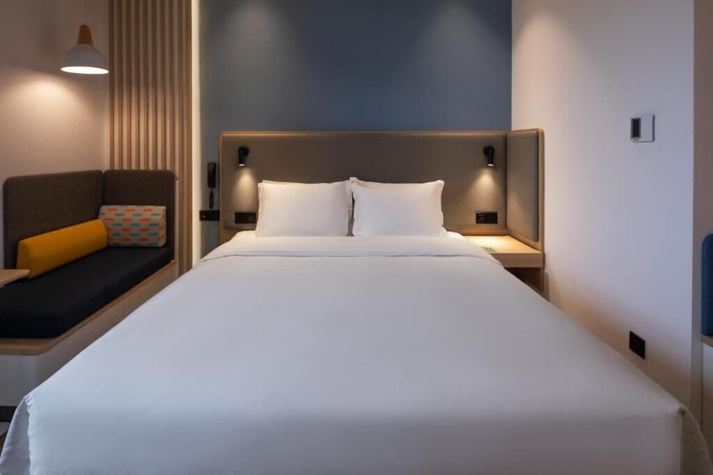 A neatly made bed with white linens, a headboard with integrated lighting, a bedside table with a lamp, and a cushioned bench with a colorful pillow.