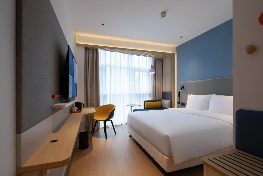 A modern hotel room with a large bed, a desk with a chair, a TV, and a window with curtains.