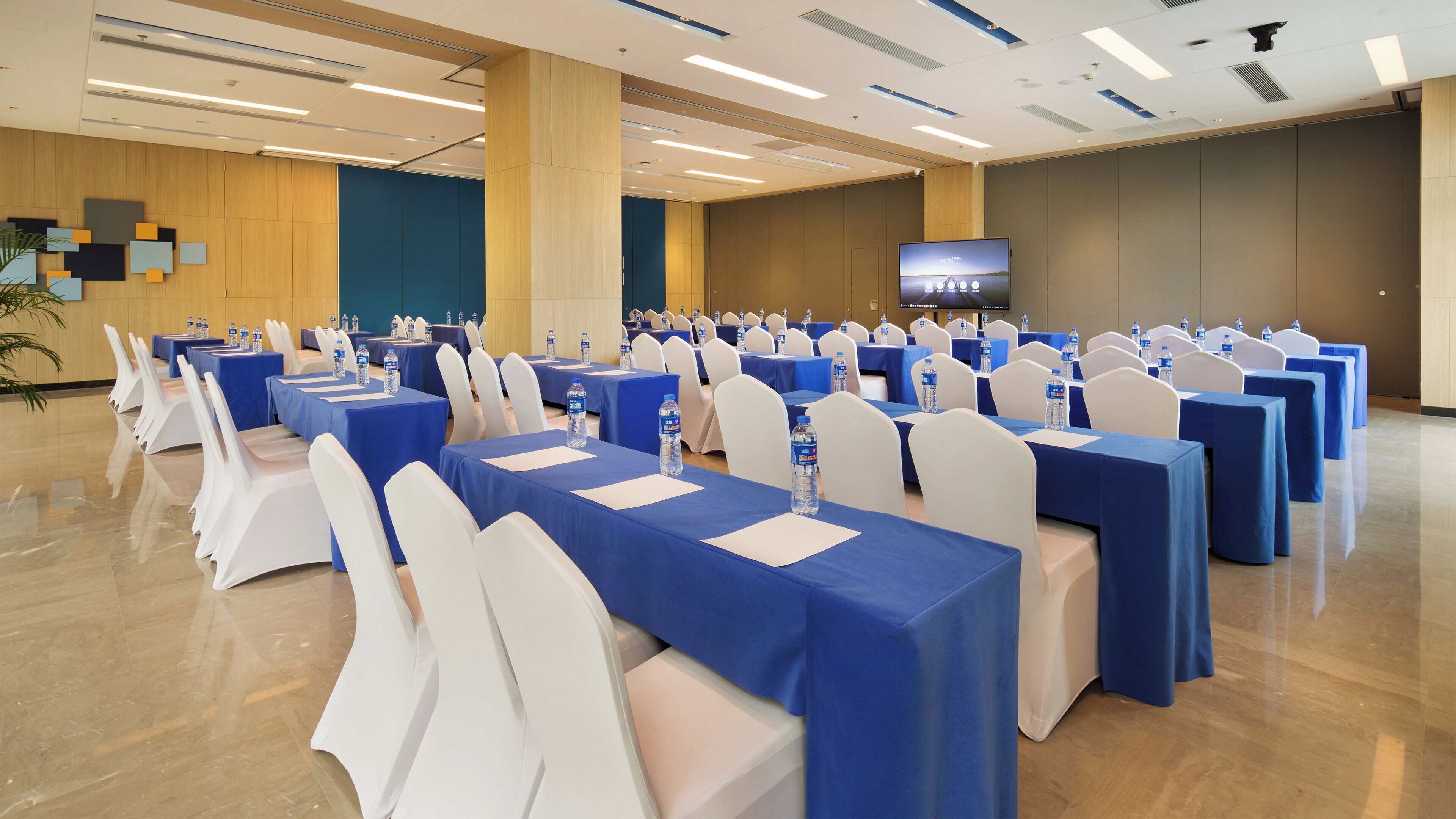Una sala conferenze con file di tavoli, sedie e uno schermo televisivo.