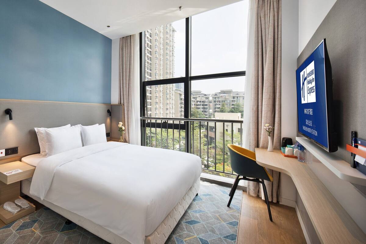 A hotel room with a large bed, a desk with a chair, a TV, and a window with a city view.