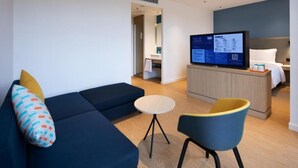 A modern hotel room with a blue sofa, a small round table, a yellow chair, a TV on a wooden cabinet, and a visible bathroom in the background. - Holiday Inn Express Shenzhen North Station by IHG (Shenzhen)