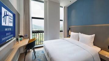 A hotel room with a bed, a desk, a chair, a nightstand, a phone, and a view of the outdoors.