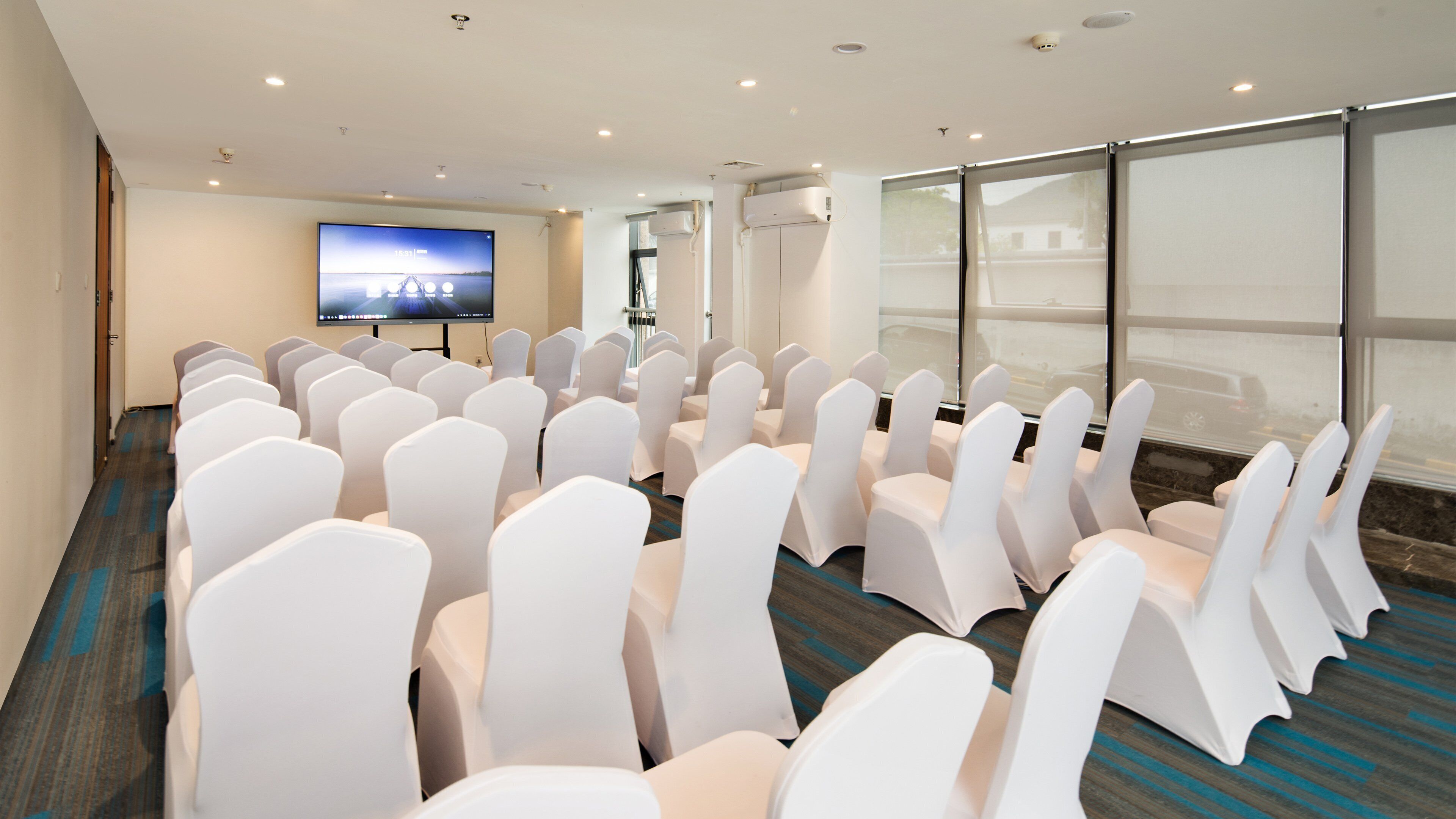 A conference room with rows of white chairs, a large screen, and a view of the outdoors through large windows.